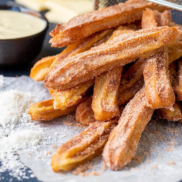 Golden-brown churros coated in cinnamon sugar, with a side of white dipping sauce and coconut flakes.
