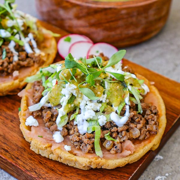 Two sopes with ground meat, refried beans, lettuce, cheese, salsa verde, and crema on a wooden board.