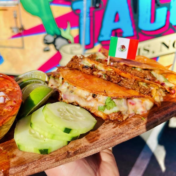 Cheesy birria tacos with a Mexican flag pick, limes, cucumbers, and consommé on a wooden board.